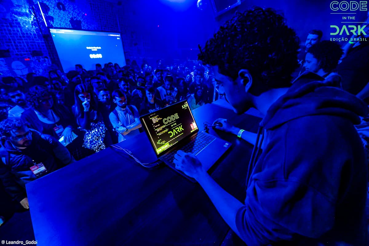 Erick Nathan, a boy with curly hair, wearing a dark blue sweatshirt, in front of a computer with programming codes and a lot of spectators around him, the photograph contains a bluish tone from the lights of the event in the dark