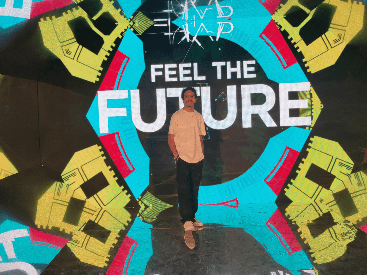 Erick Nathan, a boy with curly hair, wearing a white shirt, black pants and white sneakers in front of a mirrored LED panel reading "Feel the future"